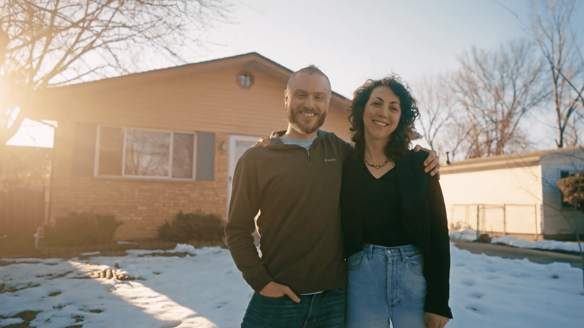 Two people smile at the camera while standing side-by-side on the snow-covered yard in front of their home.