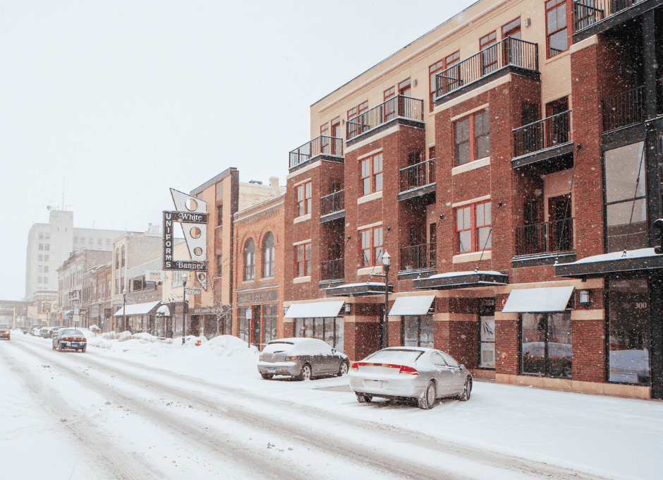 Snowy street lined with brown buildings and parked snow covered cars